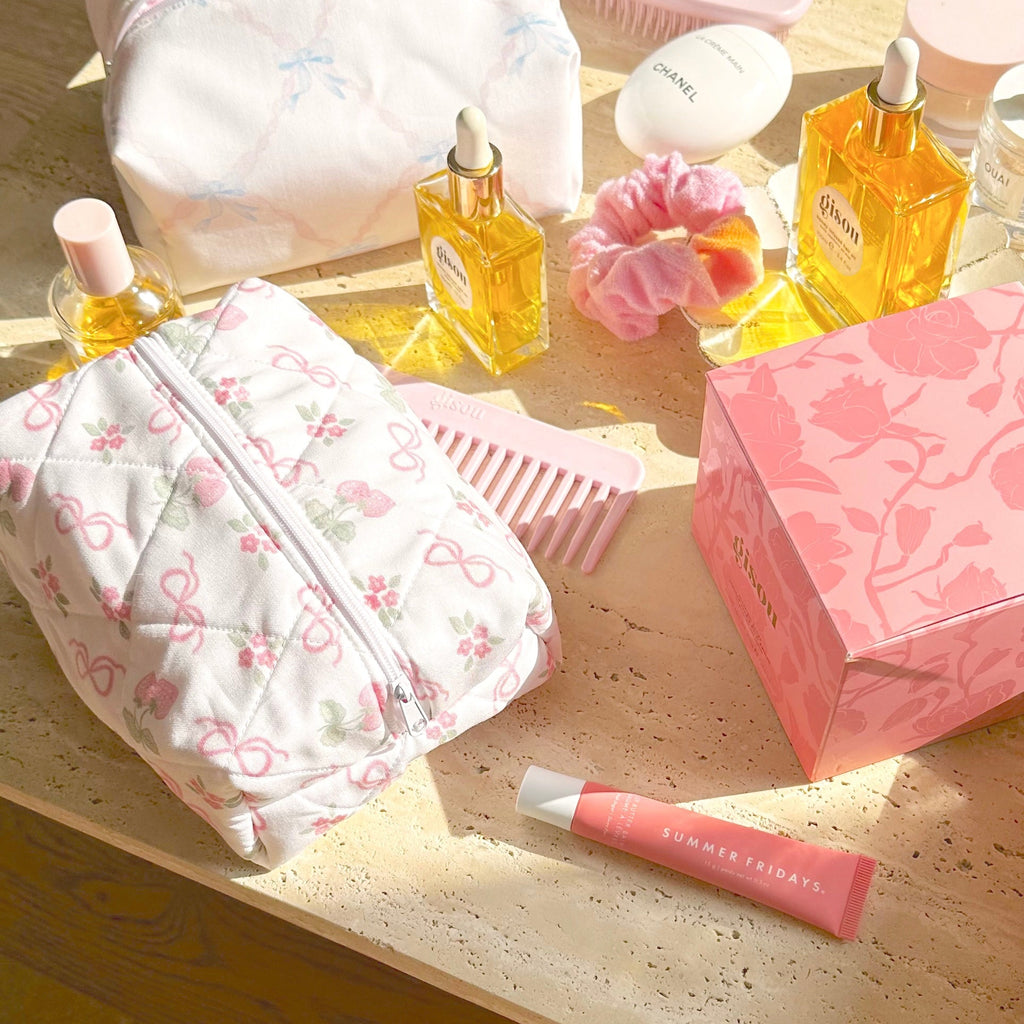 White bow-and-flower zip pouch with pink trim, on table with beauty bottles and brush