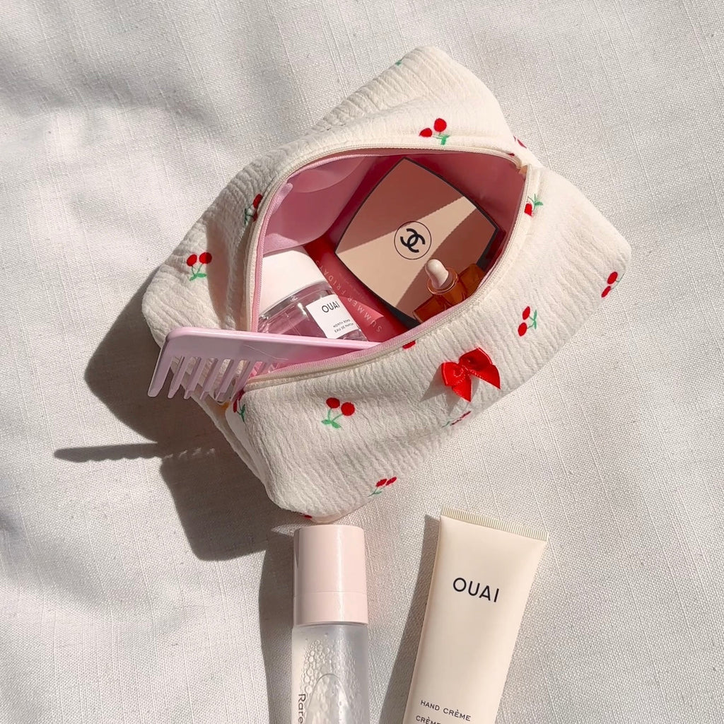 White zip pouch open with cherry embroidery and red bow, holding beauty items