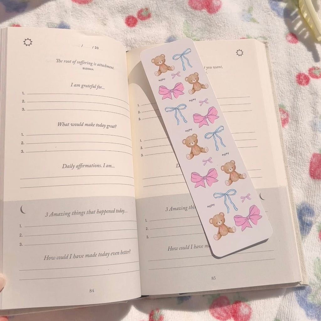Paper bookmark with teddy bears, pink bows, and blue ribbon bows on a light background.