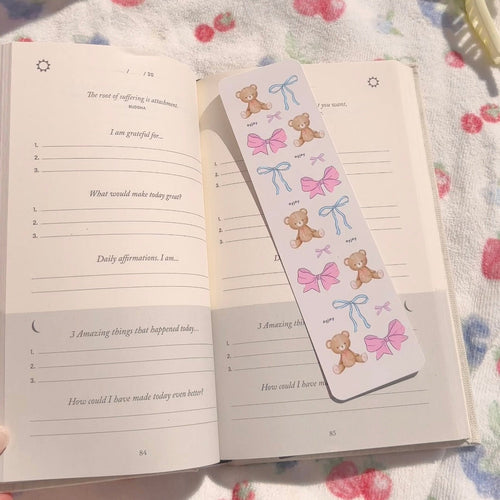 Paper bookmark with teddy bears, pink bows, and blue ribbon bows on a light background.