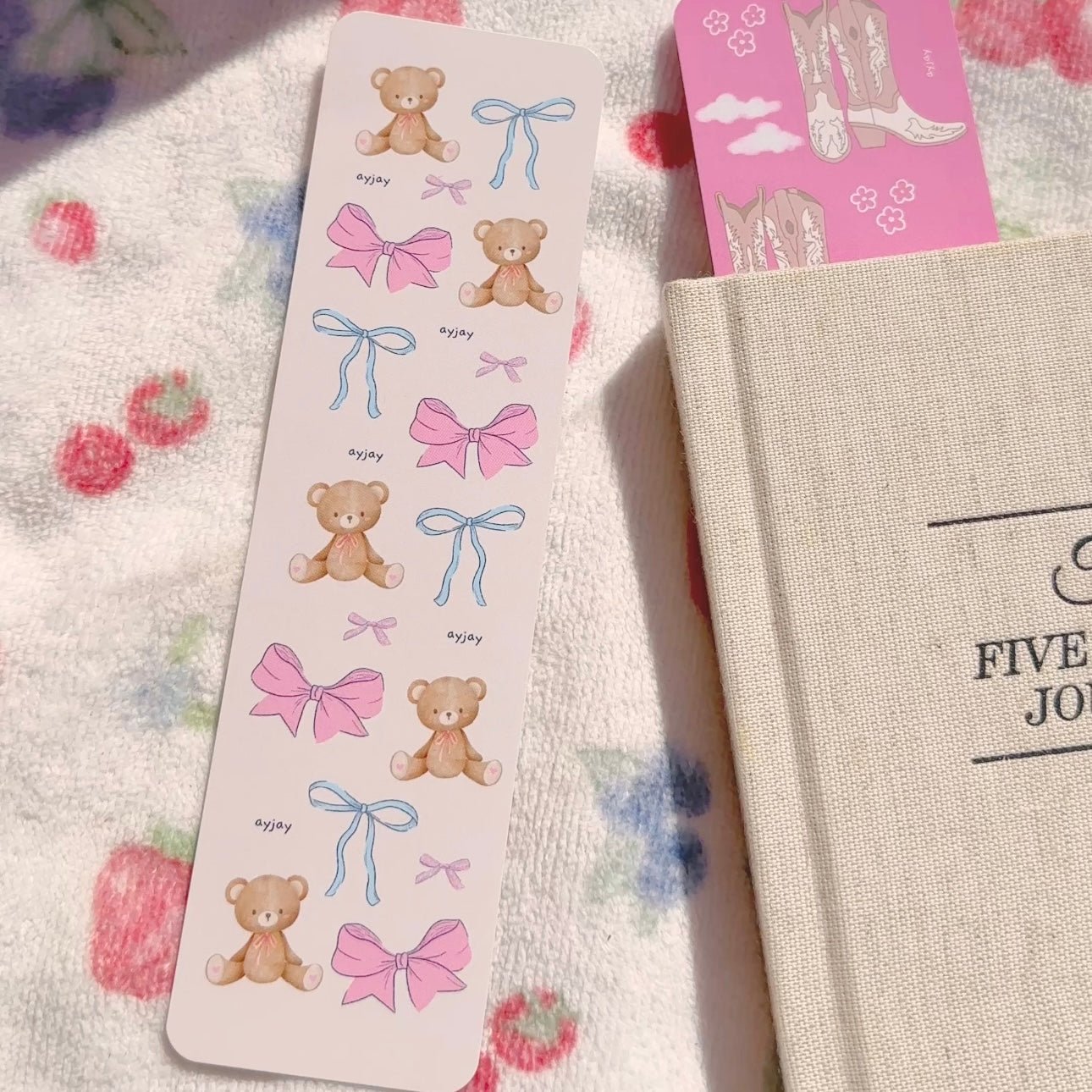 Tall paper bookmark card with teddy bears and pink bow graphics, seen beside a notebook.