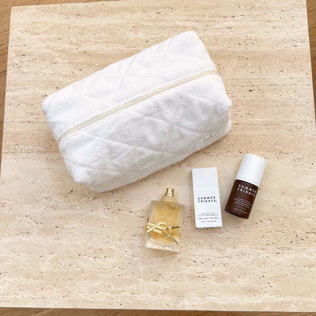 White terrycloth zip pouch, closed, with beauty bottles and cards beside it